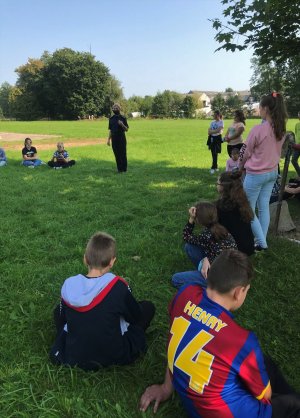 Policjantka stoi przed grupą dzieci i młodzieży, zebranych na terenie przy szkole. Część z nich siedzi na trawie