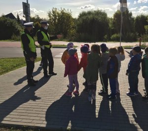 Dwóch policjantów wydziału ruchu drogowego stoi z grupą przedszkolaków przed przejściem dla pieszych