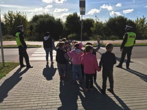 Dwóch policjantów ruchu drogowego stoi z grupa przedszkolaków przed przejściem dla pieszych