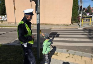Policjant wydziału ruchu drogowego stoi z dzieckiem przy przejściu dla pieszych. Udziela mu wskazówek, co zrobić, aby światło zmieniło się na zielone
