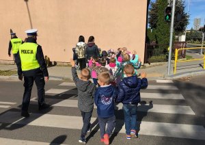 Policjanci wydziału ruchu drogowego przechodzą z dziećmi przez przejście dla pieszych. Na sygnalizatorze świeci się światło zielone