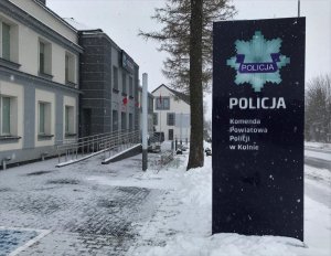 Na zdjęciu granatowa tablica, na której umieszczona jest policyjna odznaka oraz napis:  Policja Komenda Powiatowa Policji w Kolnie. W tle budynek Komendy Powiatowej Policji w Kolnie.