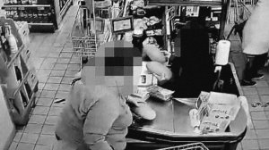 Zrzut ekranu z monitoringu sklepowego obejmujący rejon kas. Przy kasie kobieta  jedna ręką pakuje zakupy do koszyka, natomiast drugą przykrywa swoim portfelem inny portfel, który leży na ladzie.