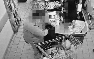 Zrzut ekranu z monitoringu sklepowego obejmujący rejon kas. Przy kasie kobieta ubrana w bluzkę chowa portfel do torebki. Obok niej stoi wózek sklepowy.