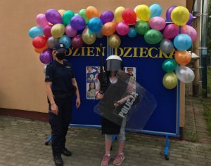Umundurowana policjanta w czarnej maseczce na twarzy stoi obok dziecka ubranego w policyjny sprzęt PZ. Za nimi niebieska tablica z napisem Dzień Dziecka oraz kolorowe balony.