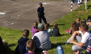 Umundurowana policjantka stoi przed grupą dzieci, które siedzą na trawie.