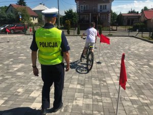 Teren przed szkołą. Świeci słońce. Umundurowany policjant Wydziału Ruchu Drogowego idzie obok dziecka jadącego rowerem i omijającego pachołki z czerwonymi flagami.
