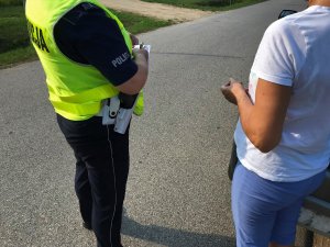 Policjanta na drodze wypisuje formularz mandatu karnego kobiecie ubranej w niebieskie spodnie i białą koszulkę z krótkim rękawem.