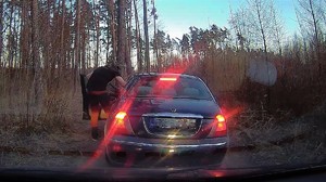 Zrzut ekranu z nagrania z kamery samochodowej. Na nim samochód osobowy stojący na drodze w lesie. Przez otwarte drzwi od strony kierowcy, do środka pojazdu  schyla się mężczyzna.