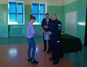 Sala gimnastyczna w szkole. Komendant Powiatowy Policji w Kolnie oraz Wójt Gminy Turośl wręczają nagrodę oraz dyplom dziewczynce.