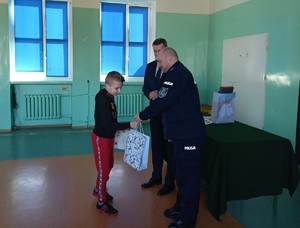 Sala gimnastyczna w szkole. Komendant Powiatowy Policji w Kolnie oraz Wójt Gminy Turośl wręczają nagrodę oraz dyplom chłopcu.