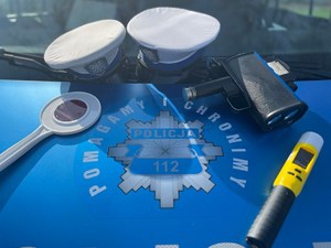 Czapki policjantów ruchu drogowego, miernik prędkości, alkomat i tarcza do zatrzymywania pojazdów leżące na masce radiowozu.