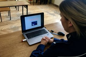 Policjantka siedzi przy biurku. Przed nią laptop. Na monitorze laptopa napis: Cyberzagrożenia i obrazek przedstawiający osobę korzystającą z laptopa.