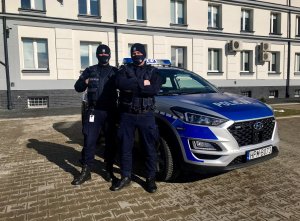 Dwóch umundurowanych policjantów stoi przy oznakowanym radiowozie. W tle budynek Komendy Powiatowej Policji w Kolnie.