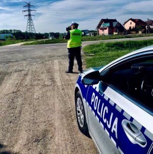 Umundurowany policjant przy drodze mierzy prędkość pojazdów ręcznym miernikiem prędkości. Obok stoi oznakowany radiowóz.