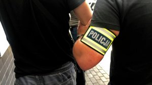 Nieumundurowany policjant, z żółtą opaską na lewym przedramieniu z napisem policja, prowadzi mężczyznę zakutego w kajdanki na ręce trzymane z przodu.