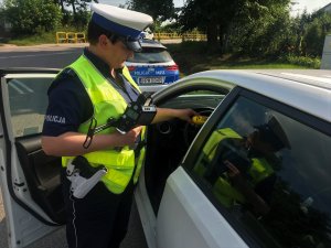Policjanta trzyma w dłoni ręczny miernik prędkości i bada stan trzeźwości kierowcy samochodu osobowego, który siedzi wewnątrz pojazdu.