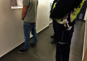 Korytarz wewnątrz budynku. Mężczyzna z kajdankami założonymi na ręce trzymane z przodu, stoi przy okienku oficera dyżurnego. Za nim stoi dwoje umundurowanych policjantów.