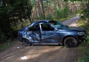 Droga w lesie. Na środku rozbity samochód osobowy w kolorze błękitnym.