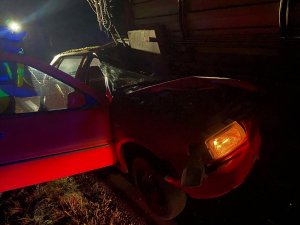 Noc. Rozbity samochód koloru czerwonego pod przyczepą rolniczą.