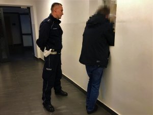 Korytarz w budynku Komendy Powiatowej Policji w Kolnie. Przy okienku stoi mężczyzna, któremu dyżurny jednostki przeprowadza badanie na zawartość alkoholu w organizmie. Za mężczyzną stoi policjant.