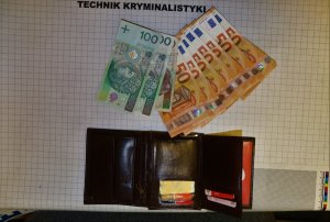 Rozłożony portfel z kartami bankomatowymi oraz dowodem osobistym. Obok portfela leżą banknoty. Na górze napis technik kryminalistyki.