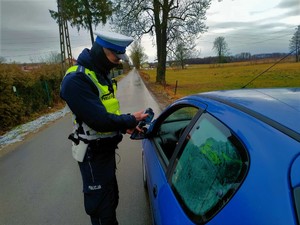 Policjant stoi przy samochodzie osobowym w kolorze niebieskim. Pokazuje na mierniku siedzącemu wewnątrz kierowcy prędkość z jaką jechał.