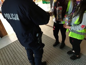 Policjant i dwie wolontariuszki WOŚP w wejściu do galerii. Policjant wrzuca do skarbonki pieniądze.