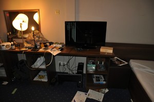 Zdemolowany pokój hotelowy. Na szafce stoi uszkodzony telewizor.