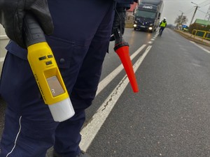 Policjant trzyma czerwoną latarkę do zatrzymywania pojazdów oraz alkomat. W tle policjantka bada stan trzeźwości kierowcy.