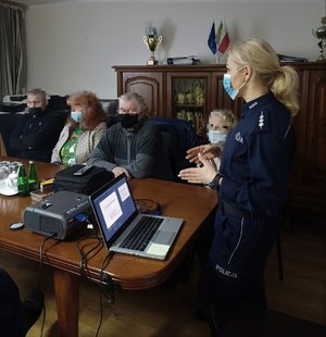 Policjantka stoi przed grupa dorosłych osób, które siedzą przy stole. Przed nią stoi laptop i rzutnik.