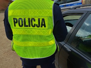 Policjant ubrany w żółtą kamizelkę odblaskową z napisem POLICJA na plecach stoi przy samochodzie osobowym w czarnym kolorze. W tle widać radiowóz.