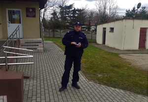 Dzielnicowy stoi na chodniku. W dłoniach trzyma notatnik służbowy