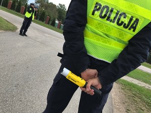 Dwoje policjantów przy drodze. Policjantka mierzy prędkość pojazdów. Policjant trzyma alkomat.