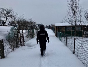 Zima. Policjant idzie ścieżką przez ogródki działkowe