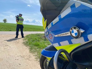 Na pierwszym planie bok policyjnego motocykla. W tle policjant przy drodze, za pomocą ręcznego miernika mierzy prędkość pojazdów.