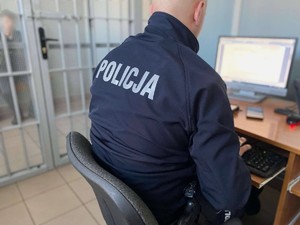 Policjant siedzący przed komputerem. W tle za kratami zatrzymany mężczyzna.