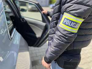 Policjant wydziału kryminalnego z opaską z napisem POLICJA na przedramieniu przy nieoznakowanym radiowozie.