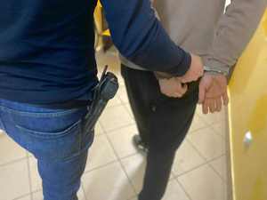 Policjant prowadzi zatrzymanego mężczyznę, który ma założone kajdanki na ręce trzymane z tyłu.