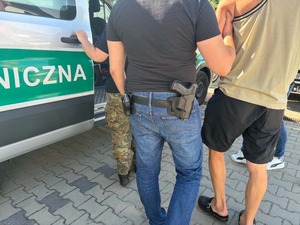 Policjant i strażnik graniczny prowadzą zatrzymanego mężczyznę do pojazdu Straży granicznej.