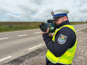Policjant przy drodze za pomocą miernika kontroluje prędkość pojazdów jadących drogą