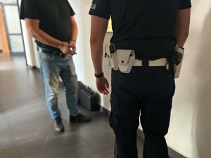 Zatrzymany mężczyzna z założonymi kajdankami na ręce trzymane z przodu i policjant na korytarzu w komendzie